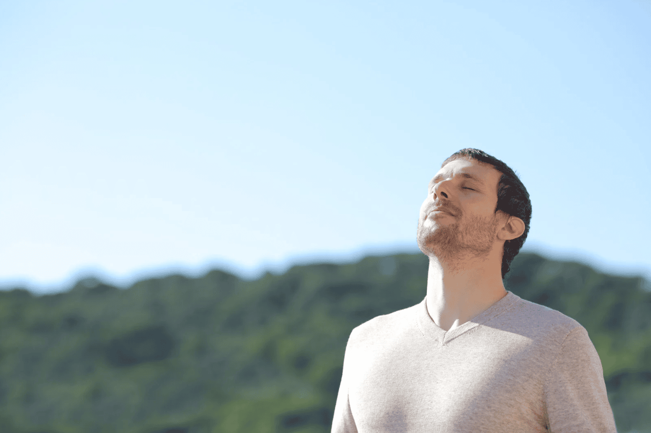 Man standing outdoors with eyes closed, taking a deep breath and enjoying the fresh air under a clear blue sky.
